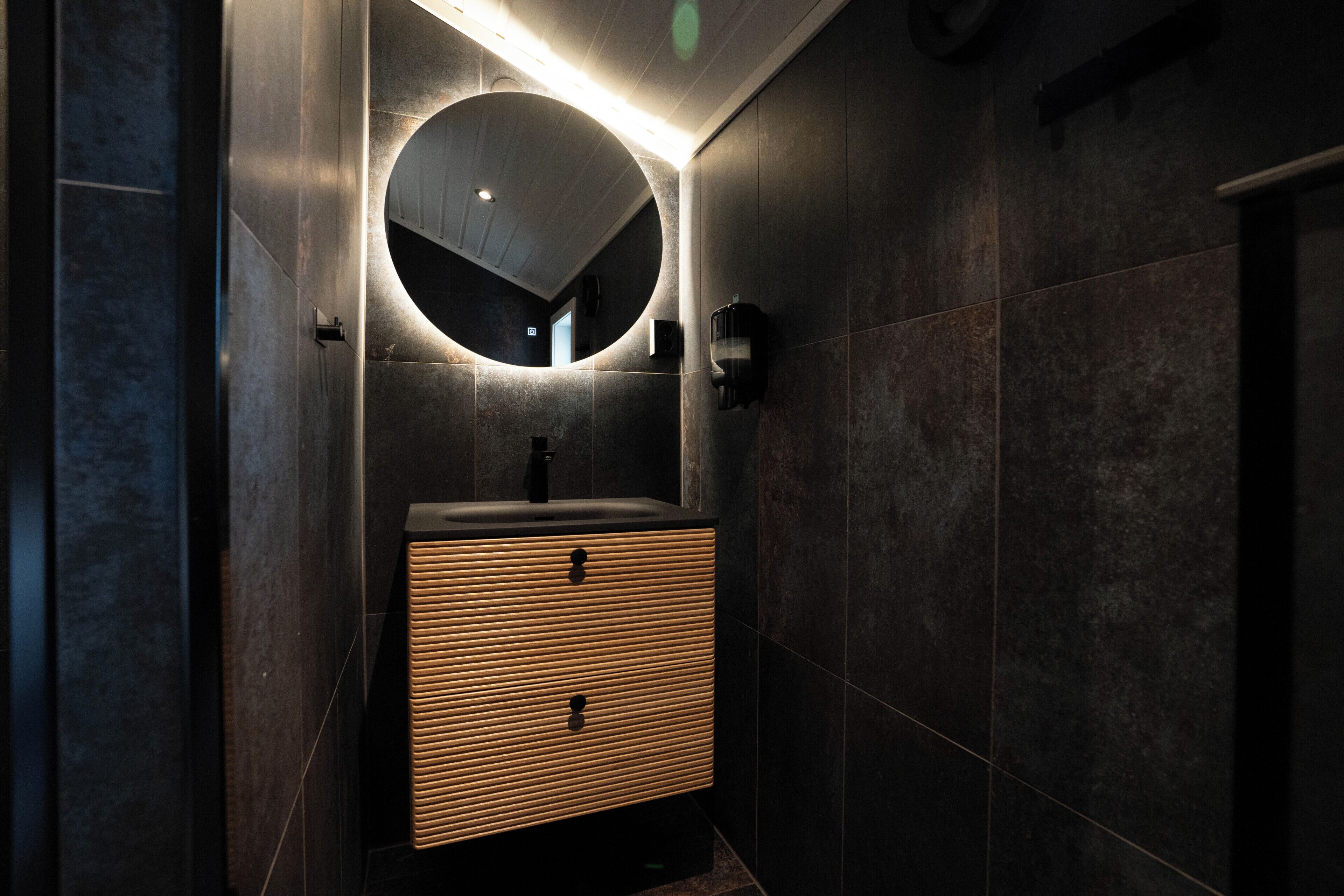 A modern bathroom with a round mirror, dark tiles, and a wooden vanity.
