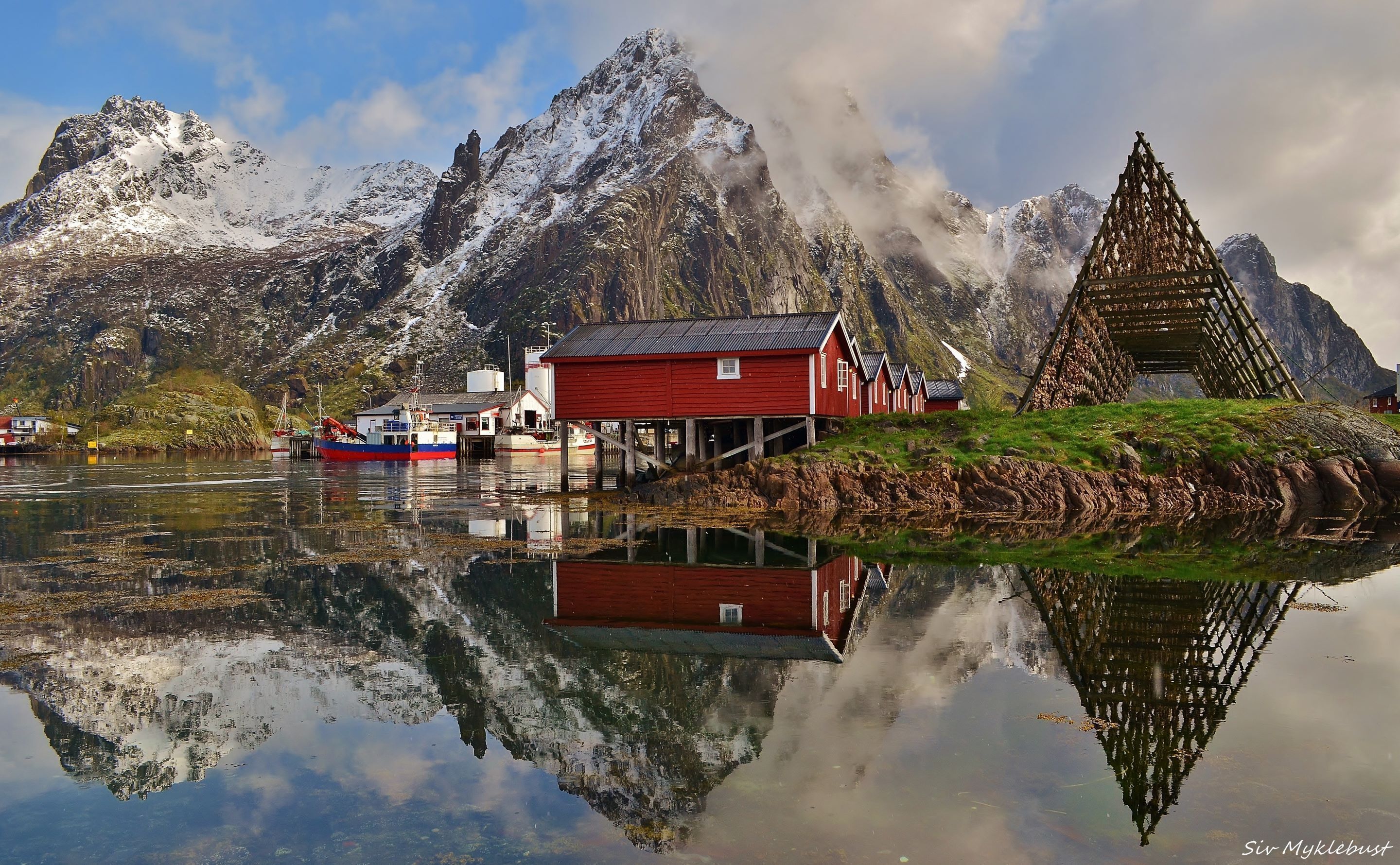 A red wooden building with a dock extending into a calm lake, surrounded by snow-capped mountains and a clear sky.