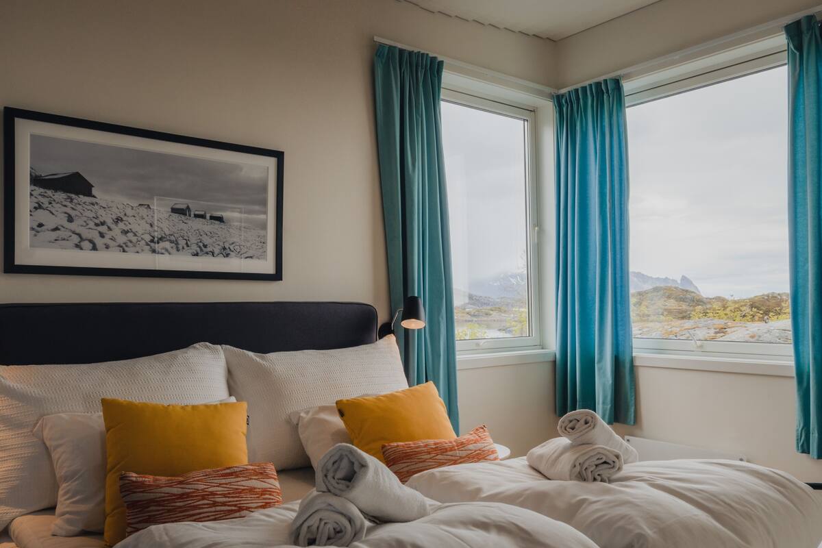 A hotel room with a bed, pillows, a headboard, a window with curtains, a framed picture on the wall, and a view of a mountainous landscape.