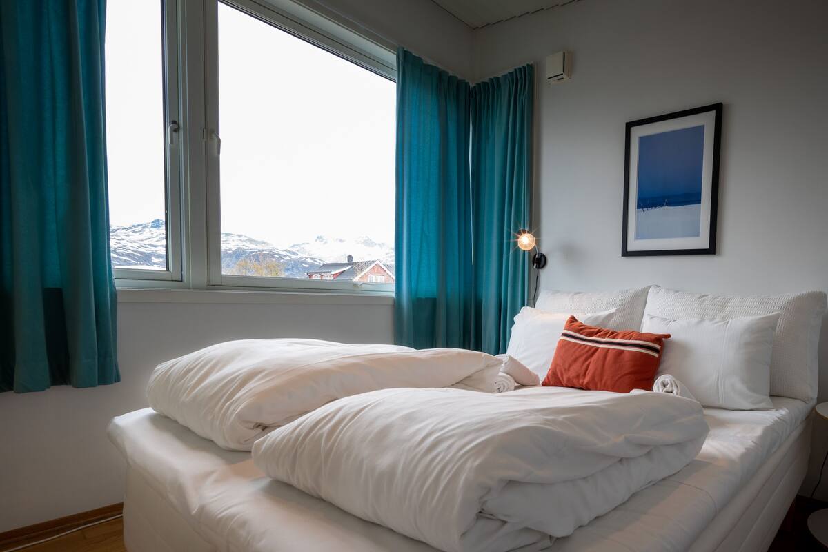 A hotel room with a bed, a window with curtains, a wall-mounted light, and a framed picture.