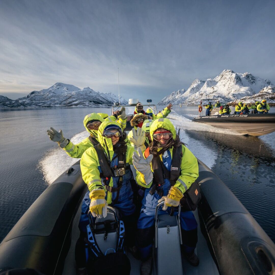 Un groupe de personnes, vêtues de tenues d’hiver jaunes et bleues, se trouve sur un bateau, avec des montagnes enneigées en arrière-plan.