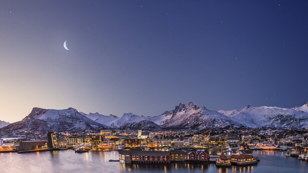 A coastal town at night with snow-covered mountains, a harbor with boats, and red houses along the waterfront. - Svinøya Rorbuer (Vagan)