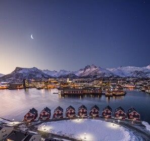 A coastal town at night with snow-covered mountains, a harbor with boats, and red houses along the waterfront.