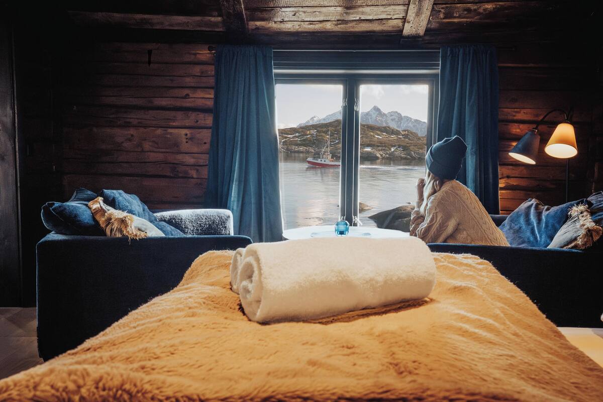A cozy cabin room with a large window, wooden walls, a bed with a rolled white towel, and a view of a boat on the water.