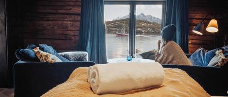 A cozy cabin room with a large window, wooden walls, a bed with a rolled white towel, and a view of a boat on the water.