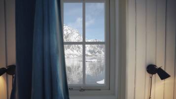 A room with a window overlooking a snowy mountain and a body of water. There are two bedside lamps and a curtain partially drawn.