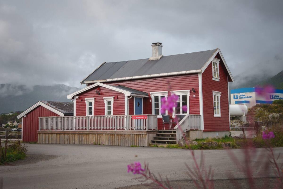 A red wooden house with a white porch and a chimney, located near a road with a mountain backdrop.