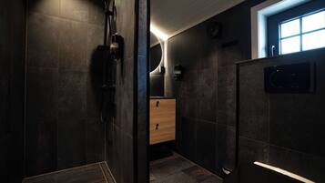 A modern bathroom with dark tiles, a wooden cabinet, a shower area, and a window providing natural light.