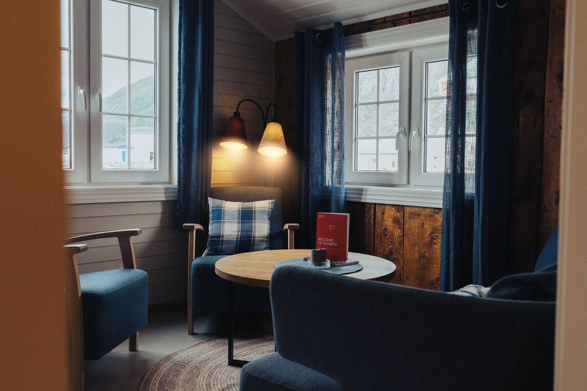 A cozy room with a round wooden table, two blue chairs, a red welcome sign, and a window with curtains.