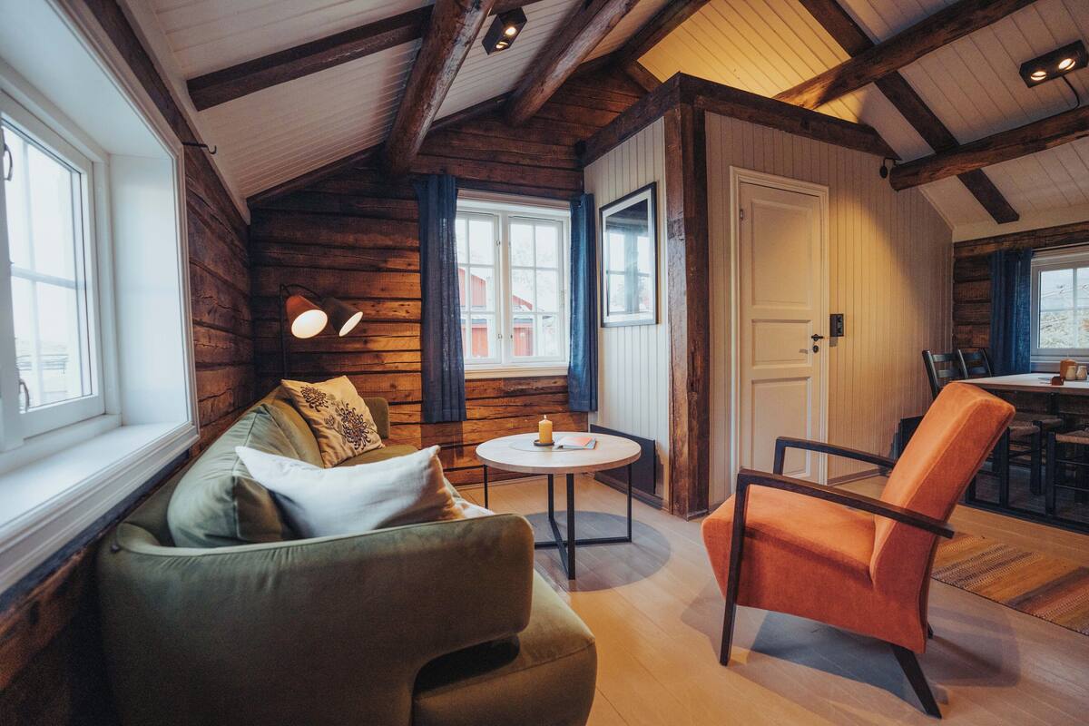 A cozy room with a wooden ceiling, a green sofa, an orange chair, a small round table, and a window with curtains.