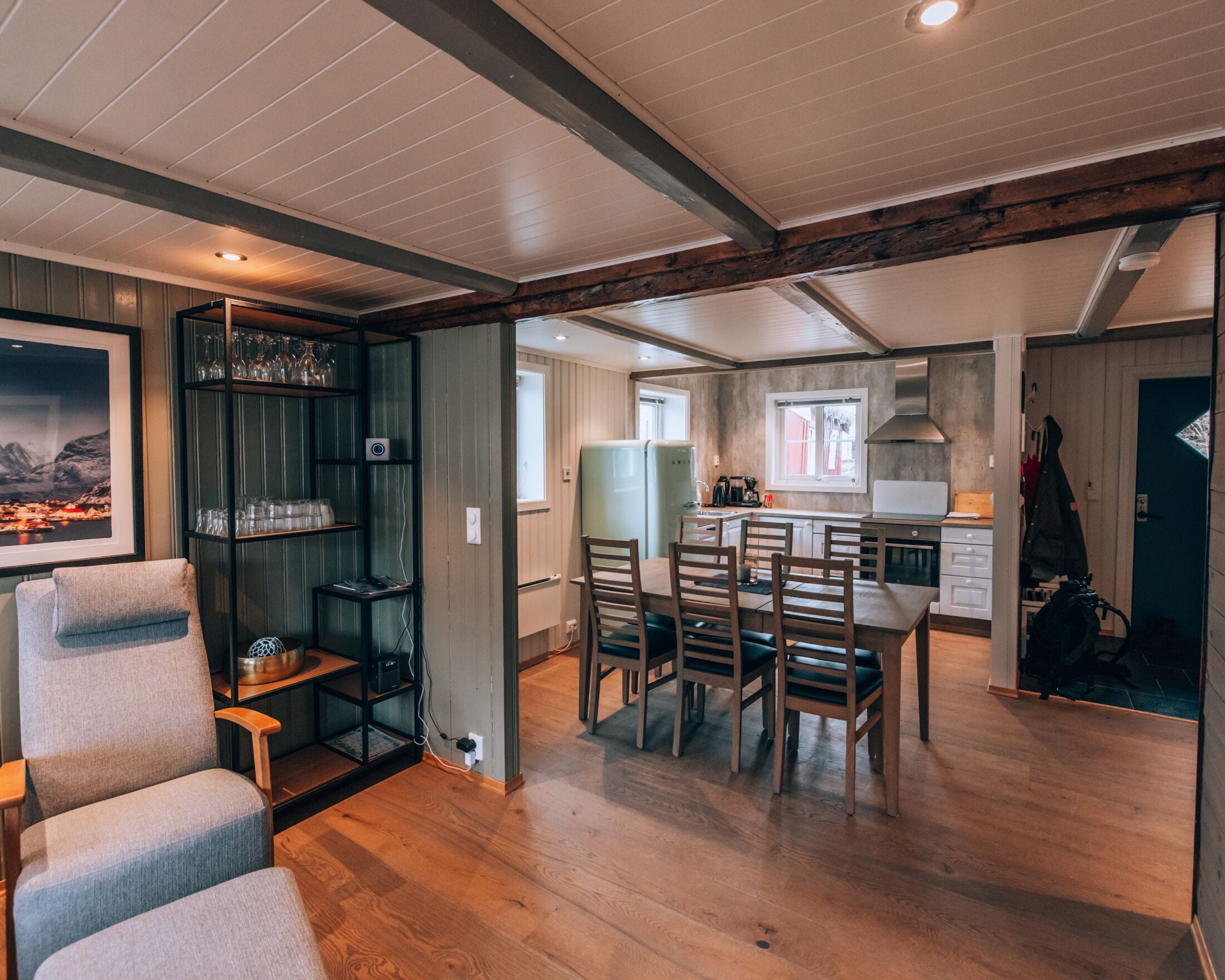 A modern kitchen with wooden cabinets, a dining area with a table and chairs, and a living space with a sofa and a bookshelf.