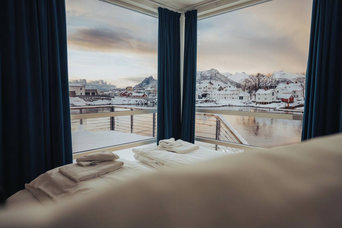 A hotel room with a bed, a window with curtains, and a view of a snowy landscape with buildings and mountains.