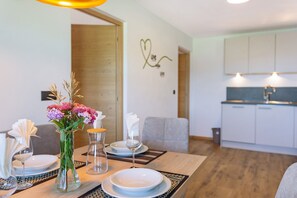 Un coin repas avec une table en bois dressée pour deux personnes, un vase rempli de fleurs et, au fond, une cuisine.