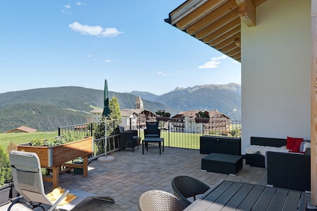 A terrace with outdoor furniture, a wooden planter, and a view of mountains and a church.