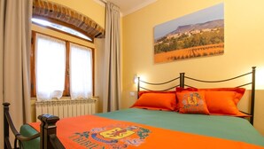A bedroom with a bed, a window with curtains, a radiator, a painting on the wall, and a bedspread with a crest. - One Bedroom Apartment (Bibbiena)