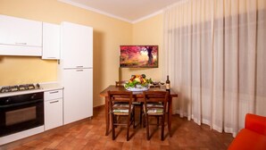A compact kitchen with white cabinets, a gas stove, and a small oven. A dining table with chairs and a fruit bowl is set against a yellow wall, with a painting above it. - One Bedroom Apartment (Bibbiena)