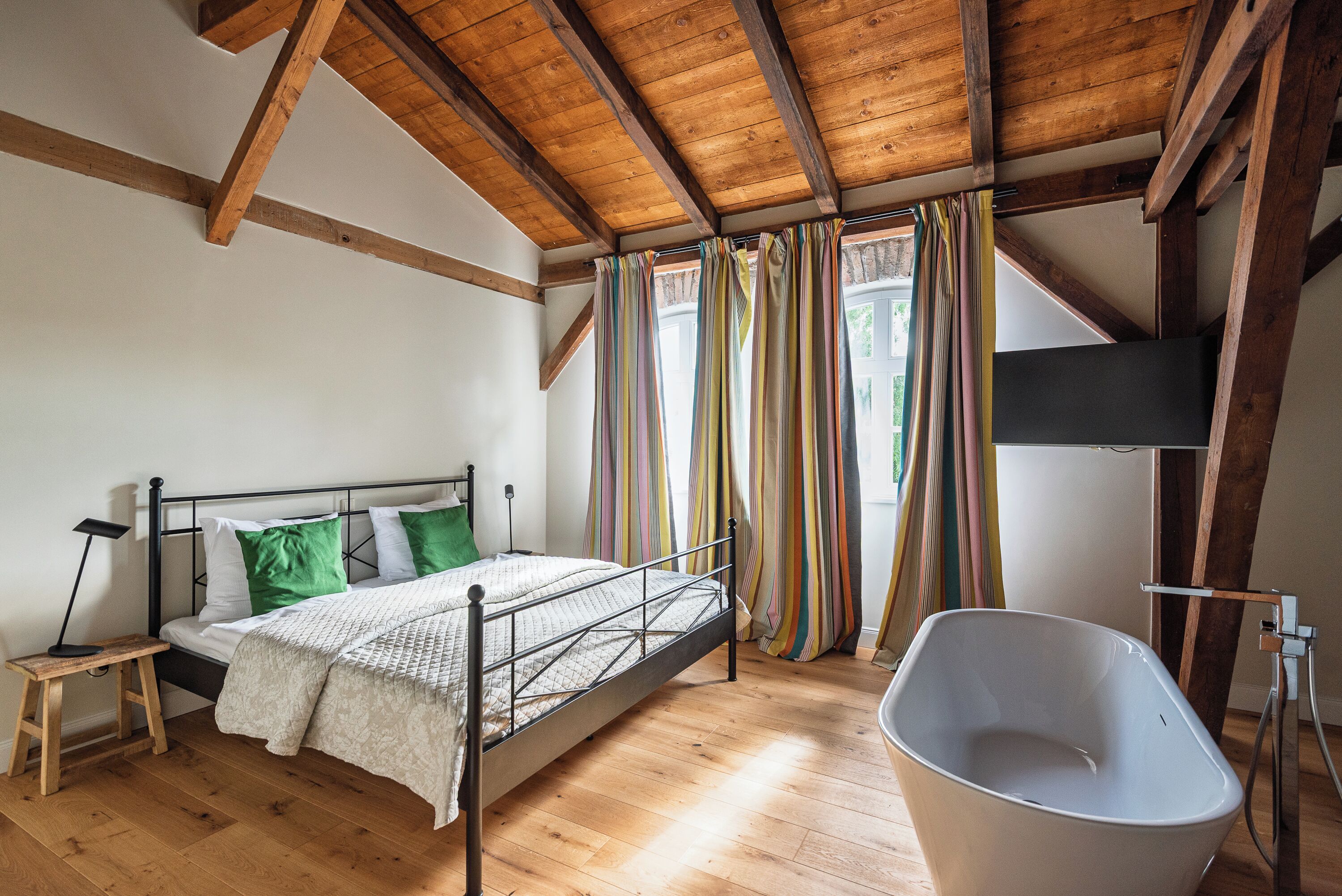 A bedroom with a wooden ceiling, a bed with a metal frame, a white bathtub, and a window with colorful curtains.