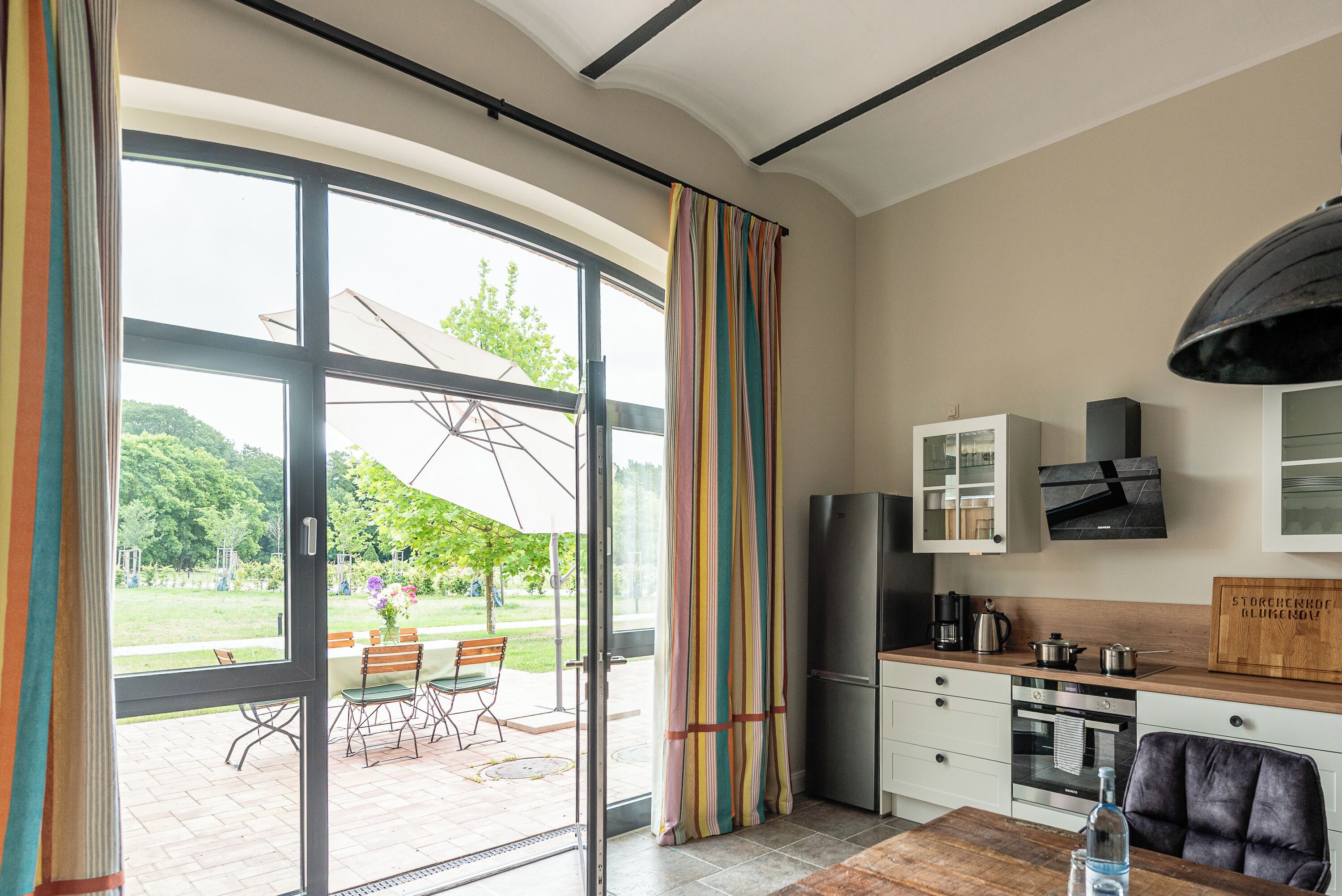 A modern kitchen with a large window, a dining area with chairs, and a view of a garden.