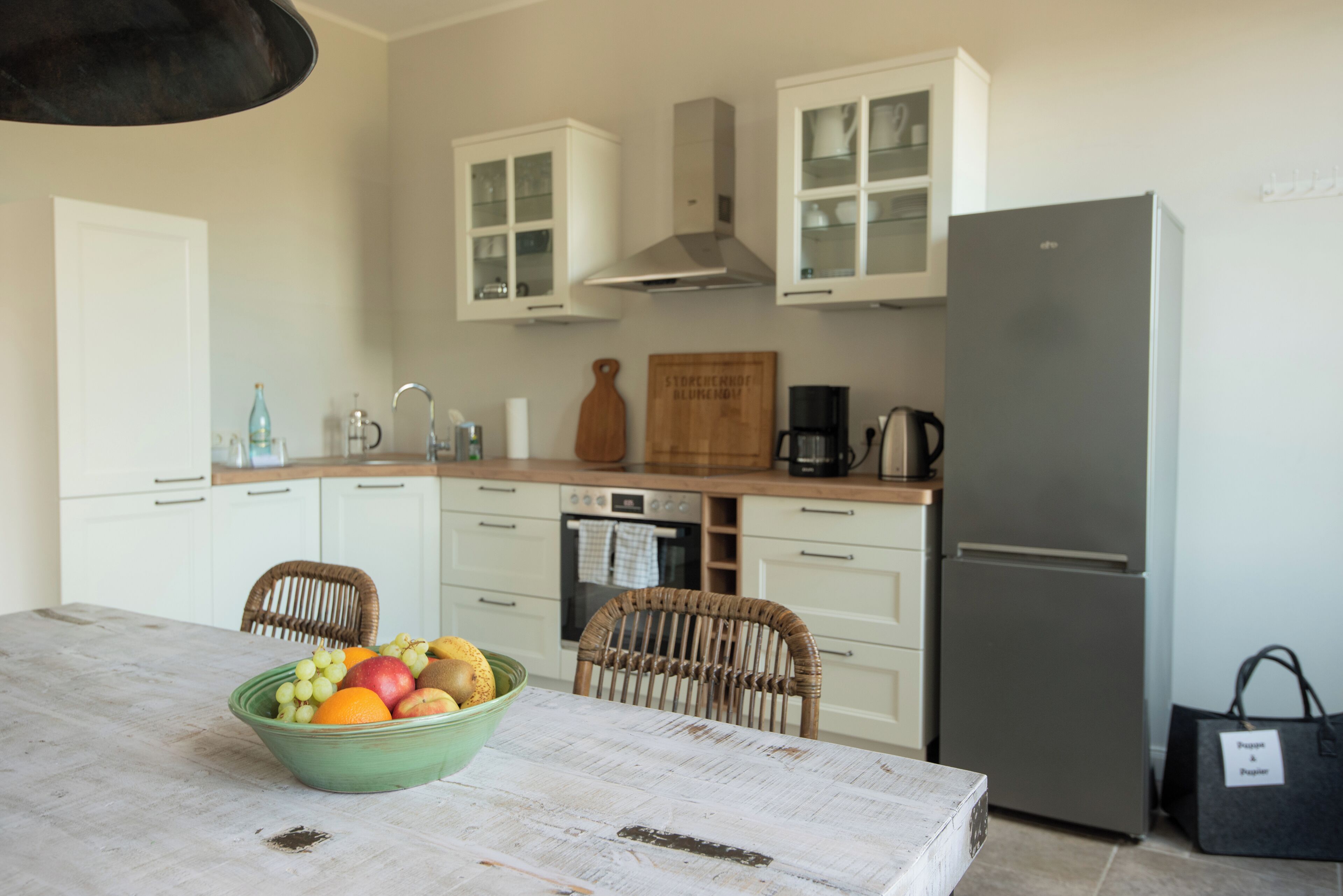 A kitchen with white cabinets, a stainless steel refrigerator, a dining table with a bowl of fruit, wicker chairs, and a coffee maker.