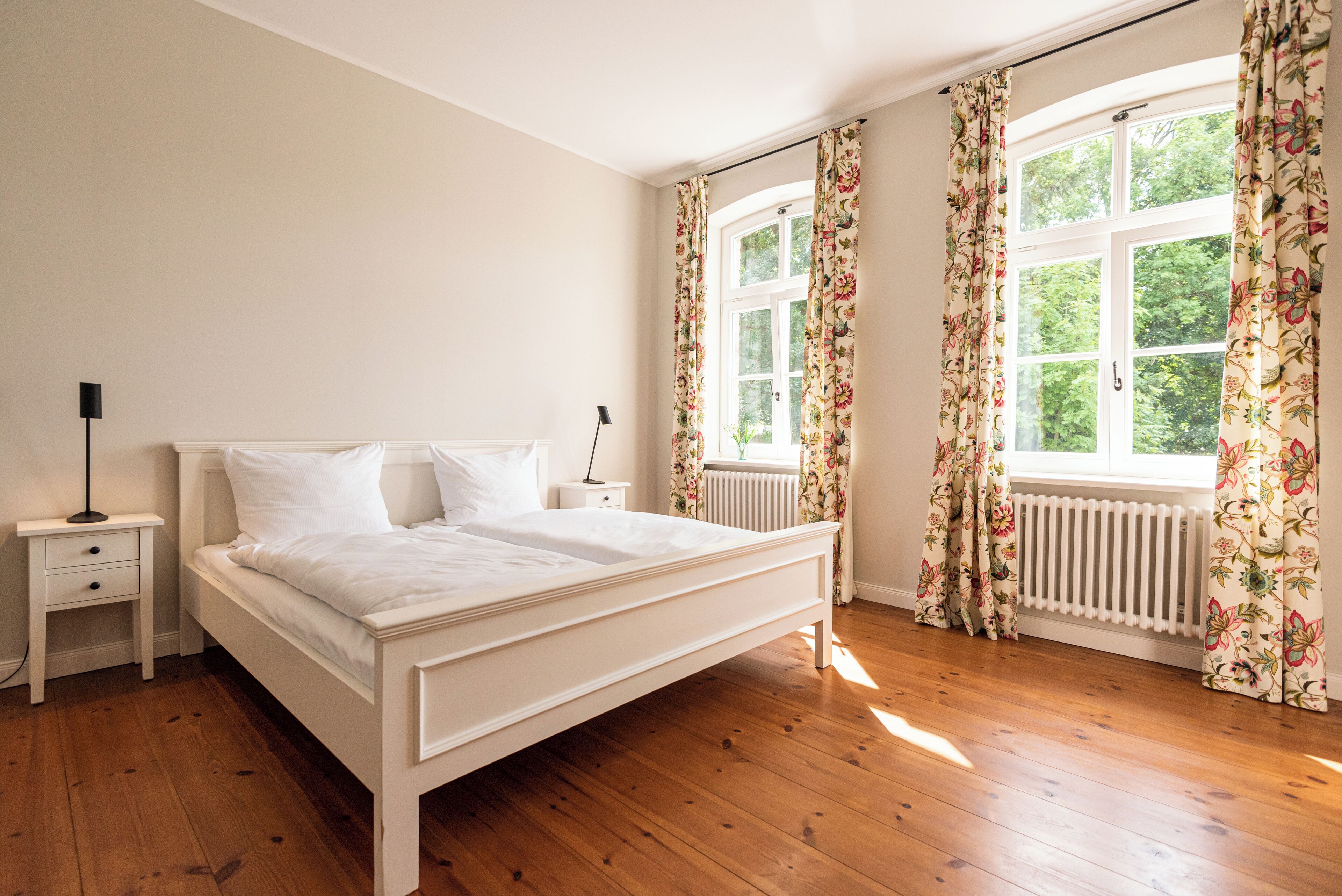 Quarto com piso de madeira, uma cama branca, mesa de cabeceira e janela com cortinas florais.