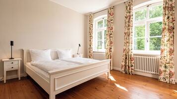 Quarto com piso de madeira, uma cama branca, mesa de cabeceira e janela com cortinas florais.