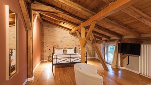 A bedroom with a sloped wooden ceiling, a brick wall, a bed, a chair, and a television.