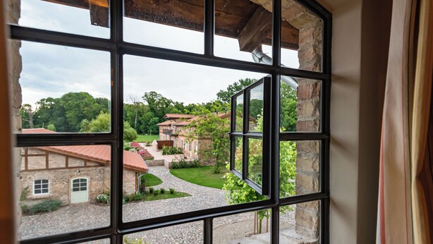 A view through a window with a stone wall and wooden beams, overlooking a courtyard with a cobblestone path, a brick building, and greenery.