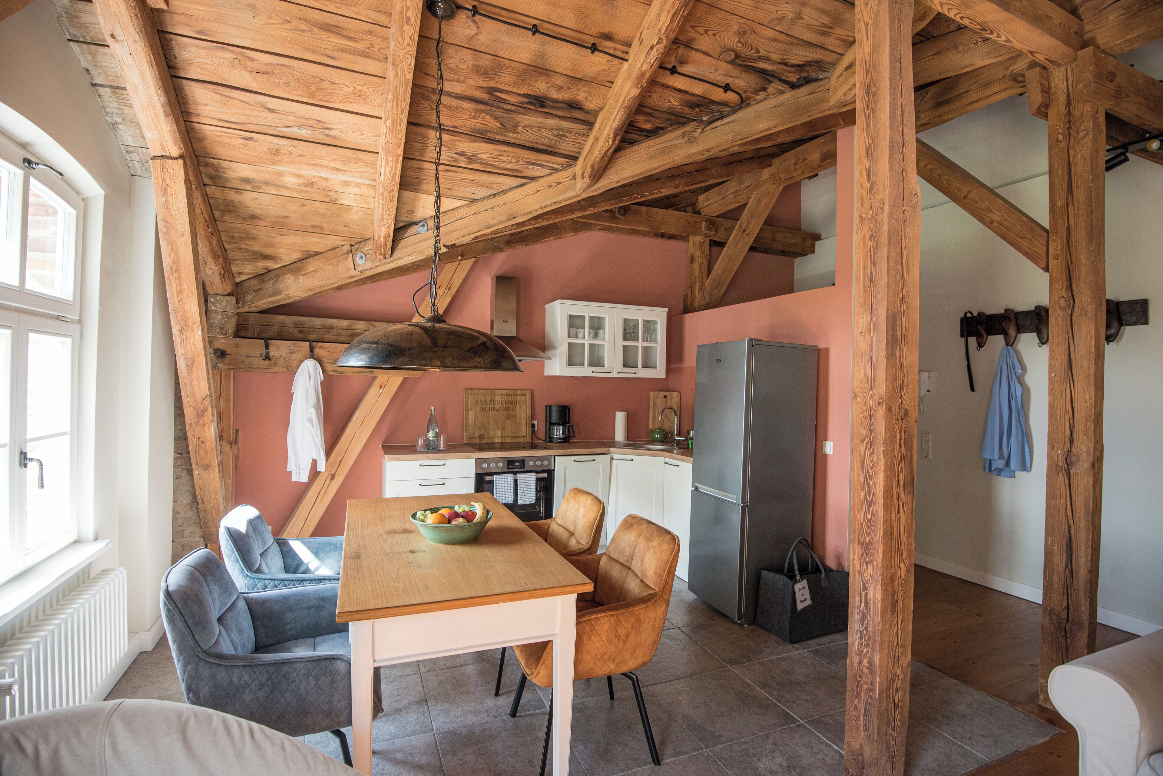 A rustic kitchen with exposed wooden beams, a dining area with a wooden table and chairs, and a pink wall.