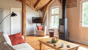 A cozy living room with a wooden ceiling, a fireplace, a television, a sofa, a coffee table, and a window with a view of greenery.