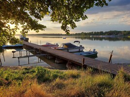Un muelle de madera con barcos, un lago tranquilo y una orilla lejana.
