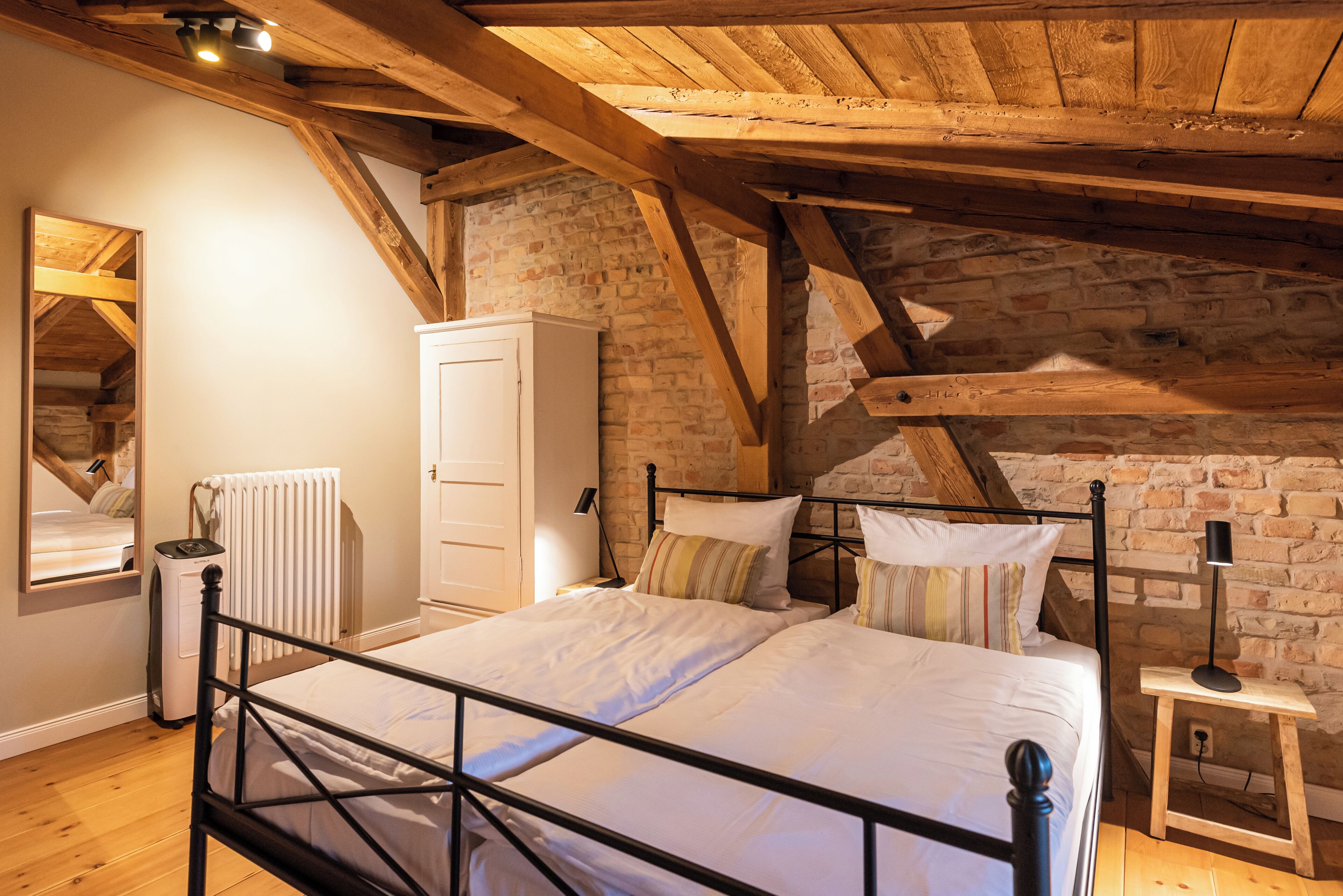 Une chambre avec un plafond en bois, un mur en briques, un lit avec une literie blanche, une armoire et une table de chevet avec une lampe.