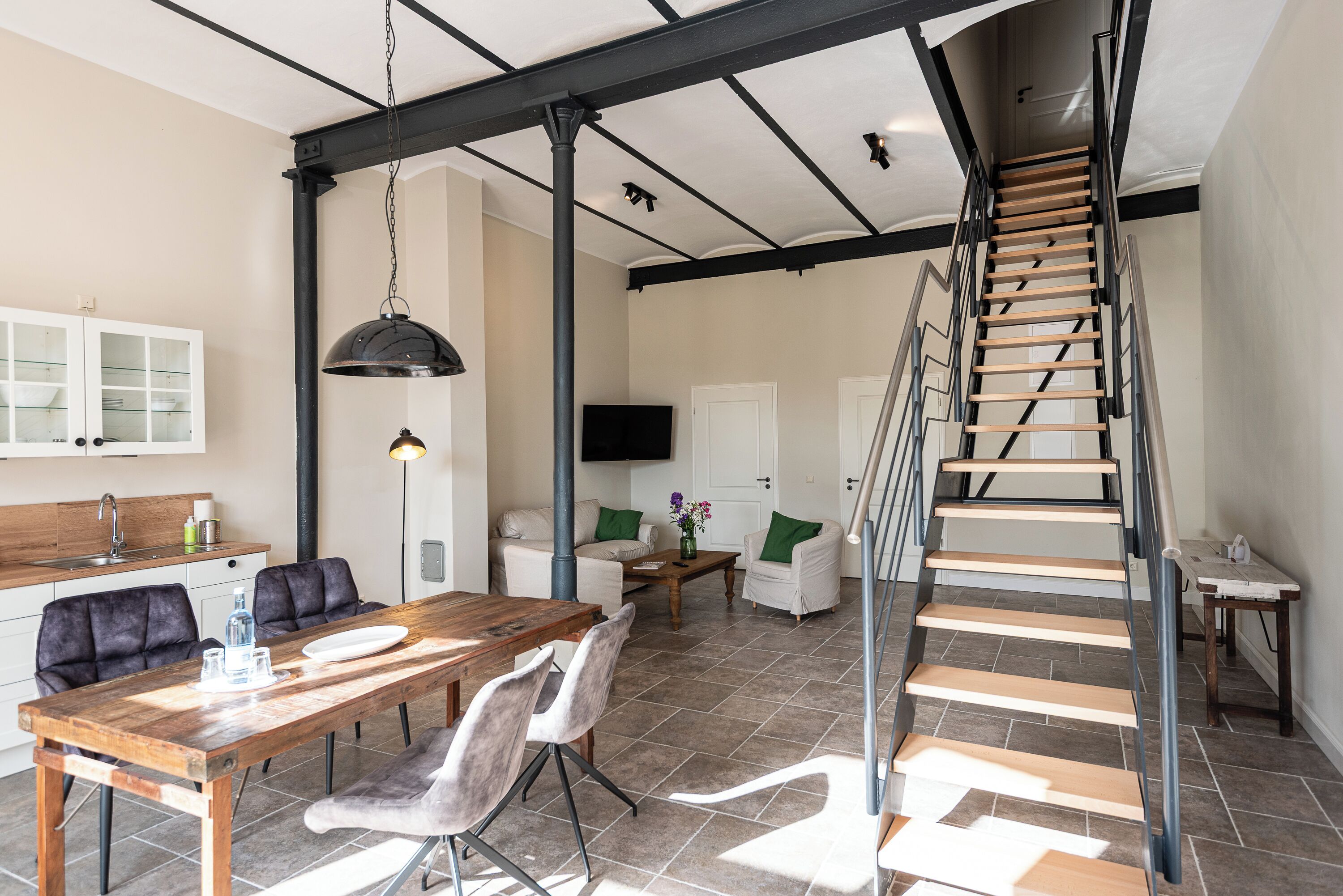 Un interior moderno con una mesa de comedor de madera, sillas, un sofá, una mesa baja y una escalera con barandilla de metal.