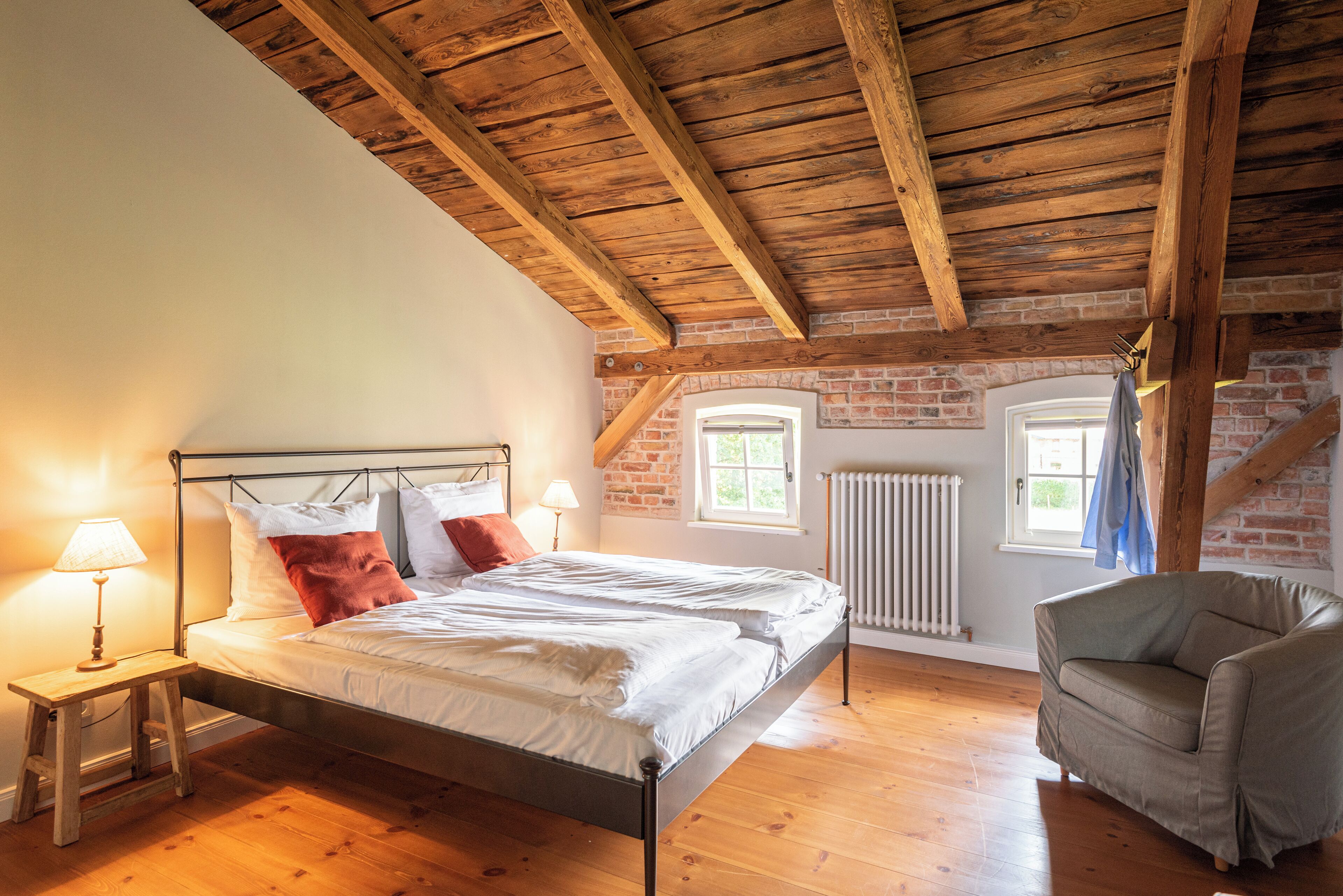 Un dormitorio con techo de madera, pared de ladrillo, cama con ropa blanca, mesita de noche con lámpara y una silla.