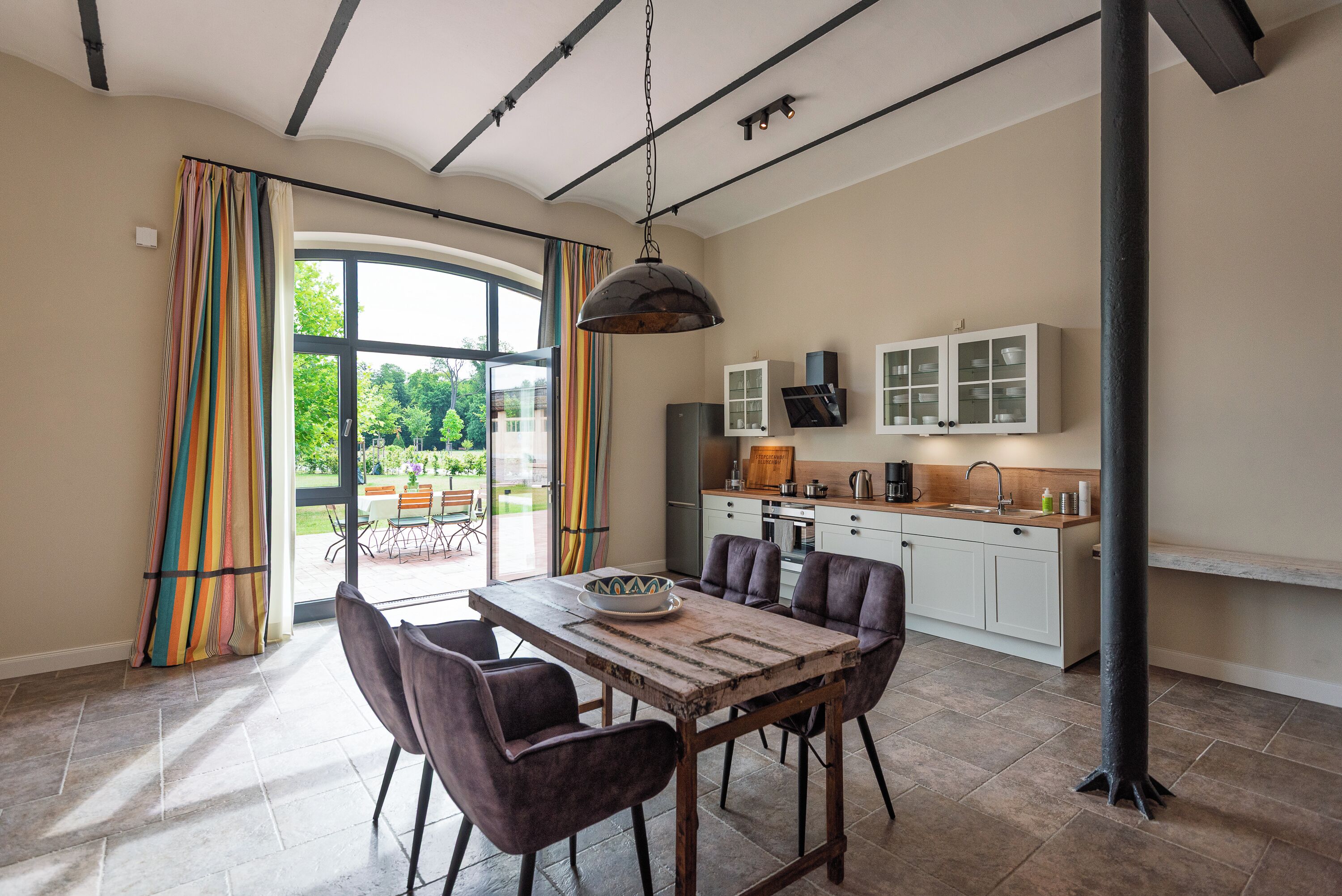 A dining area with a wooden table and chairs, a kitchen with cabinets and a sink, and a large window with curtains.