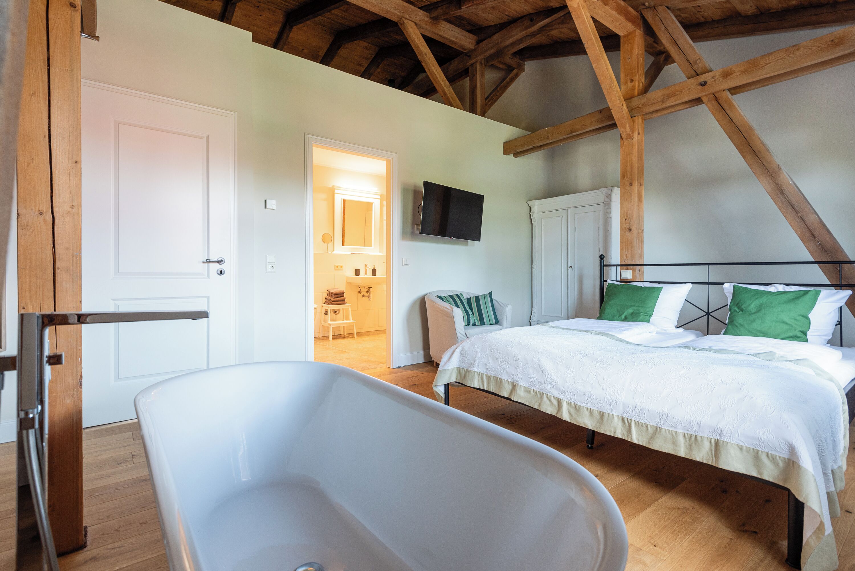 A bedroom with a large bed, a white bathtub, wooden beams, a television, and a door leading to another room.