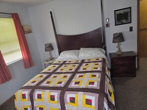 Una cama bien hecha con colcha estampada, cabecera de madera, mesita de noche con lámpara y una pequeña mesita auxiliar con un libro.