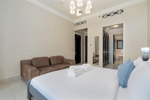 Een hotelkamer met een bed, een bruine bank, een badkamer zichtbaar door een geopende deur en een kroonluchter.