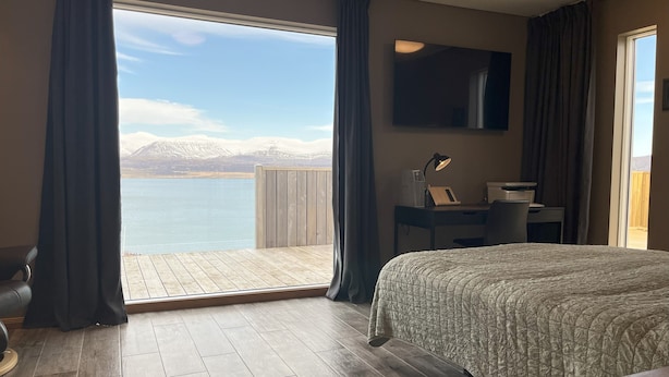 A hotel room with a bed, a desk, a chair, a TV, and a view of a lake with mountains.