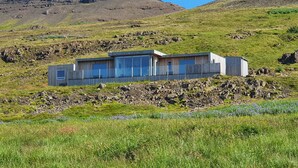 A modern building with large windows and a wooden facade, situated on a grassy hill with a rocky background.