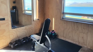 A gym with wooden walls, a bench, dumbbells, and a window with a view of a lake and mountains.