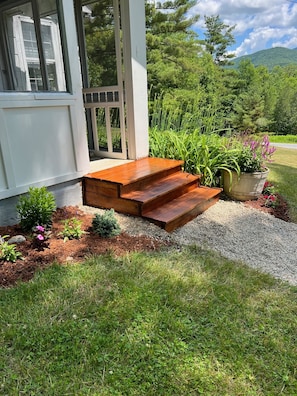 Una escalera de madera que lleva a la entrada de una casa, rodeada por un jardín con diversas plantas y flores.