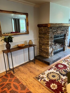 Ein Wohnzimmer mit Kamin aus Stein, einem Holzkonsole und Dekorationen, einem gemusterten Teppich und einem großen Spiegel über dem Kamin.