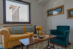 Een woonkamer met een gele bank, een blauwgroene fauteuil, een houten salontafel, een fles wijn en een schaal fruit.