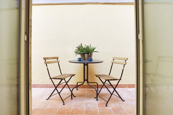 Eine kleine Terrasse mit zwei Klappstühlen und einem runden Tisch, auf dem ein Blumentopf steht, mit einer schlichten Wand im Hintergrund.