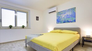 Een slaapkamer met een bed, een raam met zonwering, een airconditioner, een schilderij en een nachtkastje met een lamp.