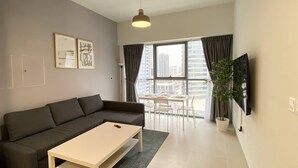Un salon moderne avec un canapé gris, une table basse blanche et un coin repas comprenant une table et des chaises. On y trouve une grande fenêtre avec des rideaux, une plante en pot et un téléviseur fixé au mur.