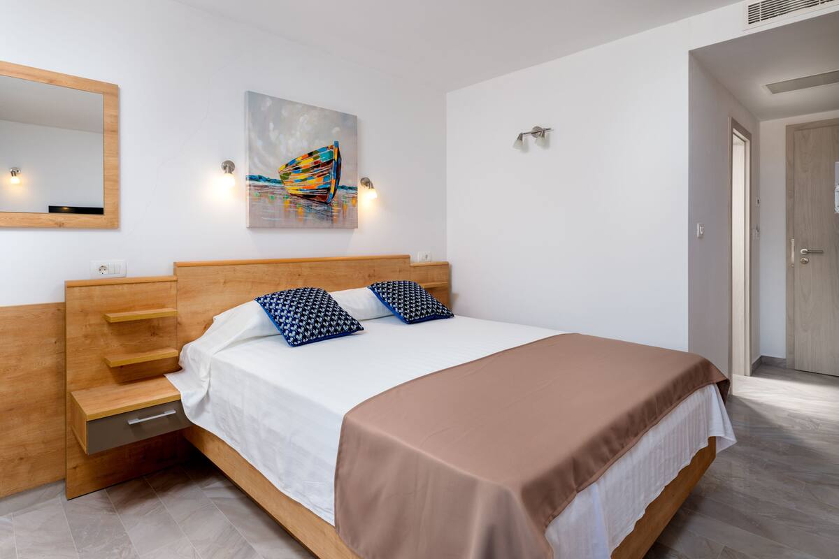 Ein modernes Schlafzimmer mit einem hölzernen Kopfteil, einem großen Bett mit brauner Decke, zwei gemusterten Kissen, einem wandmontierten Spiegel und einem Gemälde über dem Bett.