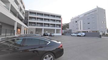 A parking lot with a black car in the foreground, a multi-story building with balconies in the background, and several white cars parked in the lot.