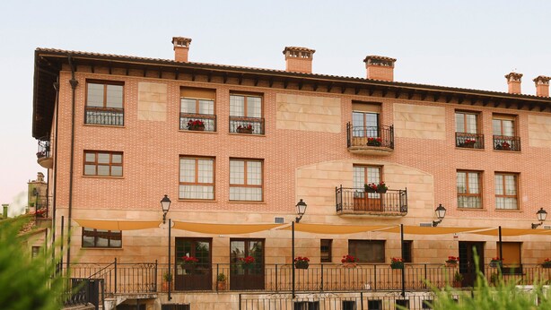 A multi-story building with a brick facade, multiple balconies with flower boxes, and outdoor seating areas with umbrellas. - Hotel Villa de Vinuesa (Vinuesa)