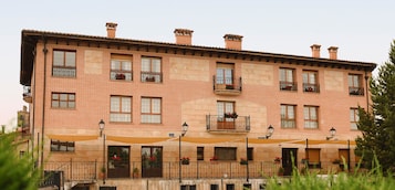 Un edificio de varios pisos con fachada de ladrillo, varios balcones con macetas de flores y zonas de estar al aire libre con sombrillas.